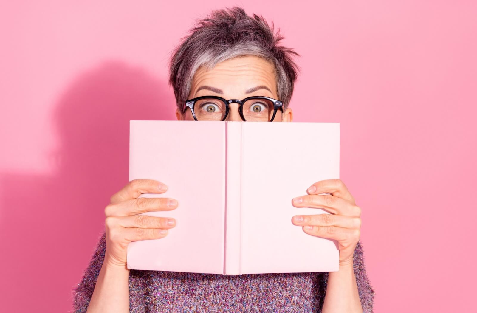 An older adult hides their face behind the cover of an open book.