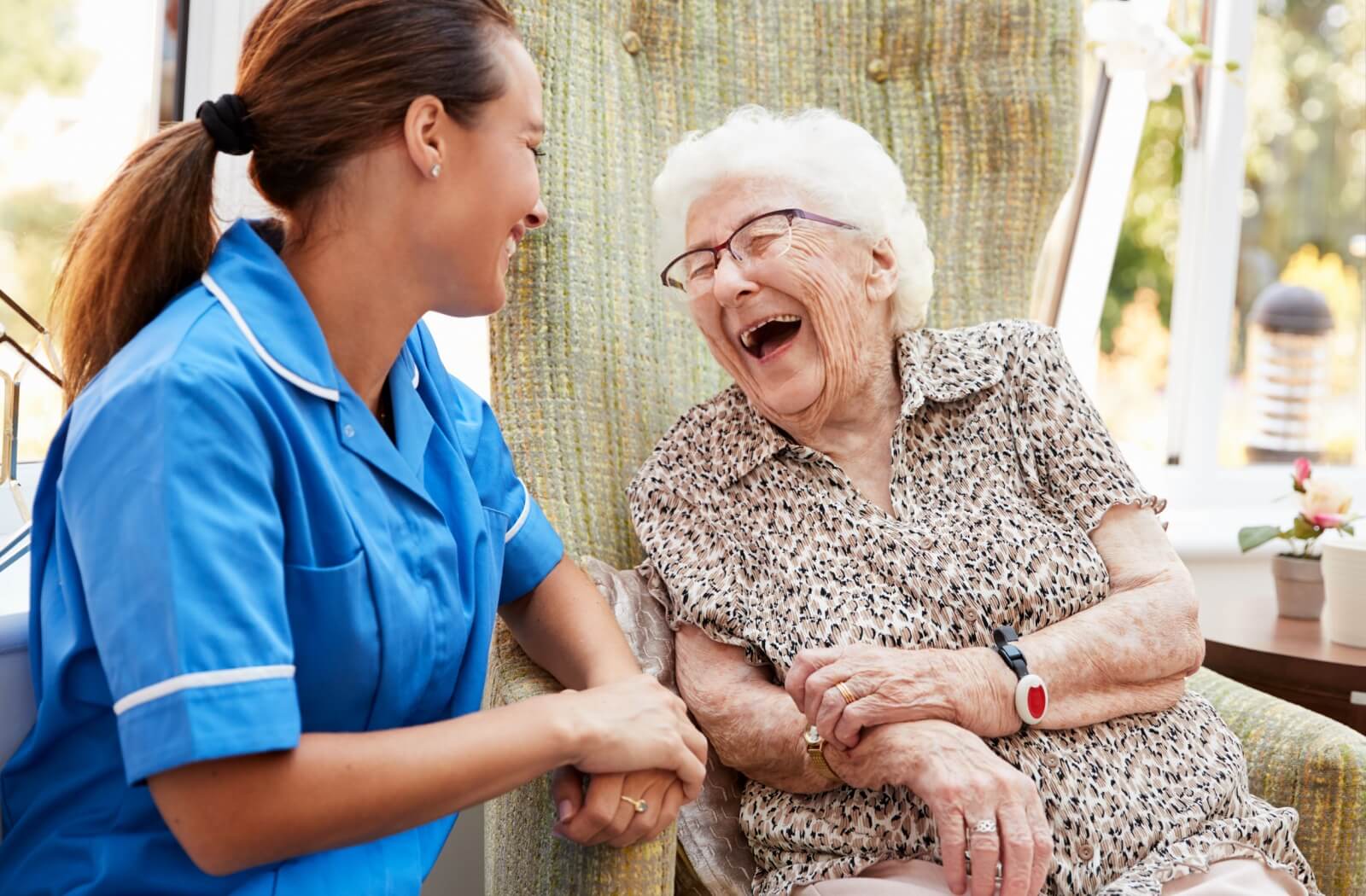 A senior in memory care sits in a comfy chair and laughs as they talk with a caregiver.