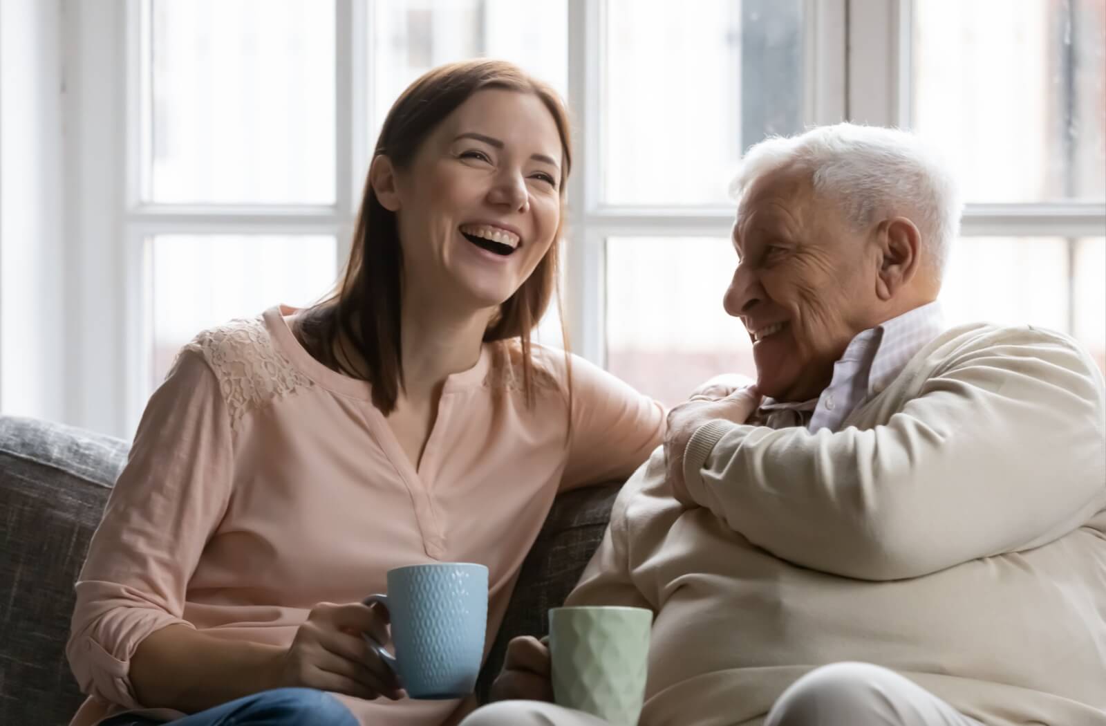 A senior with dementia sits with their adult child at home and shares a laugh.