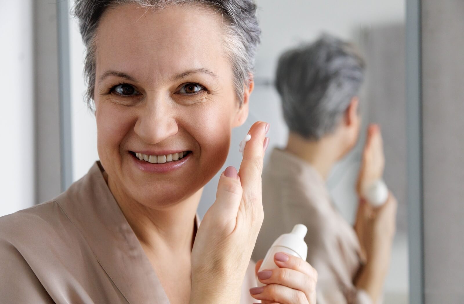 Smiling senior holding lotion bottle and applying face cream in front of mirror
