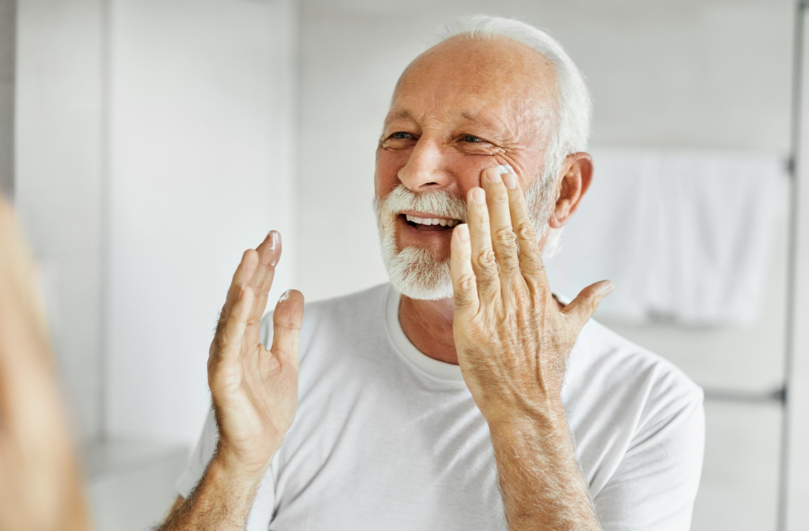 Older adult smiling while applying moisturizer to face in a bright bathroom