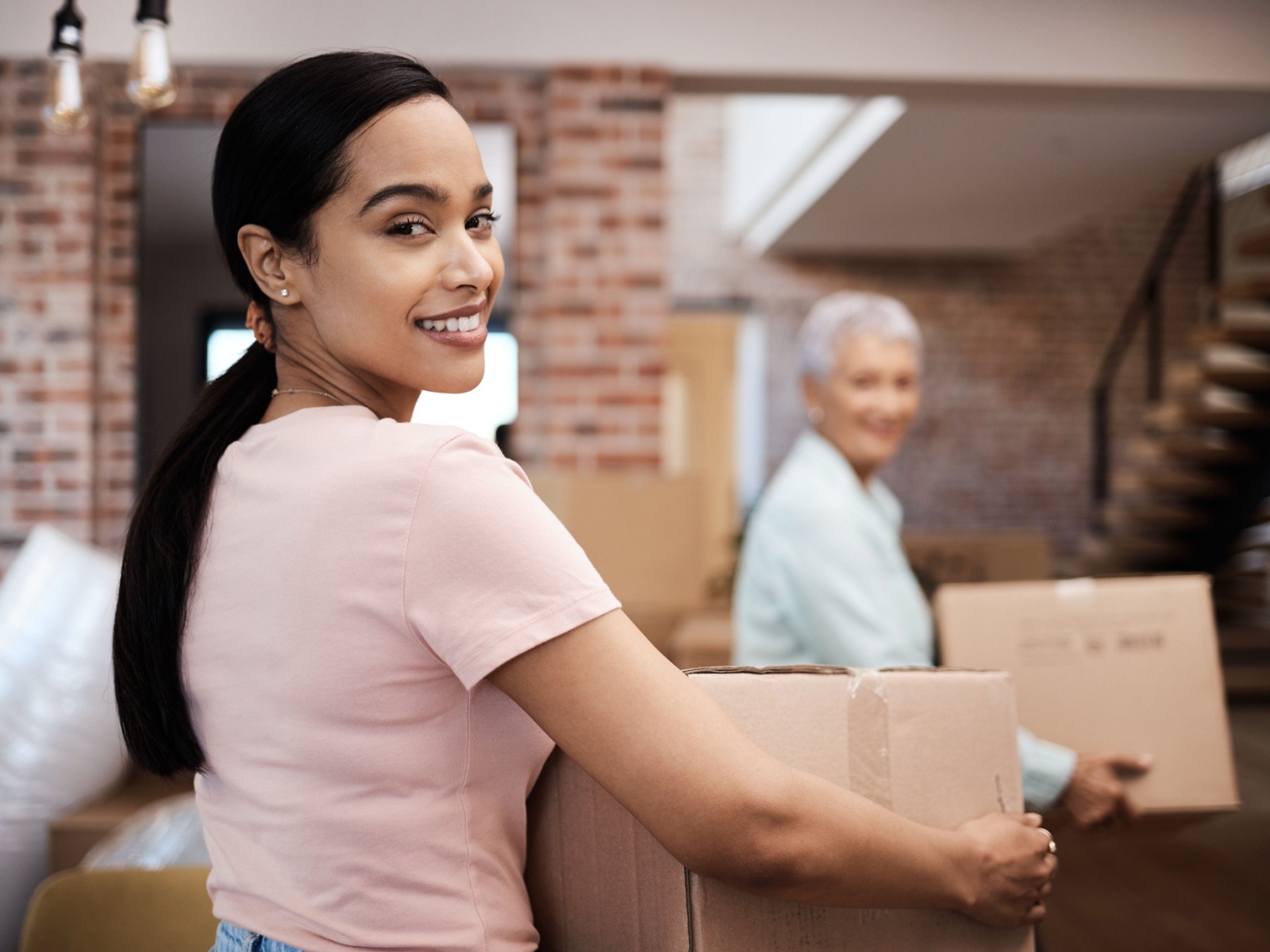 An adult child carrying a moving box smiles toward the camera as they help their senior parent pack up their belongings to move into senior living.
