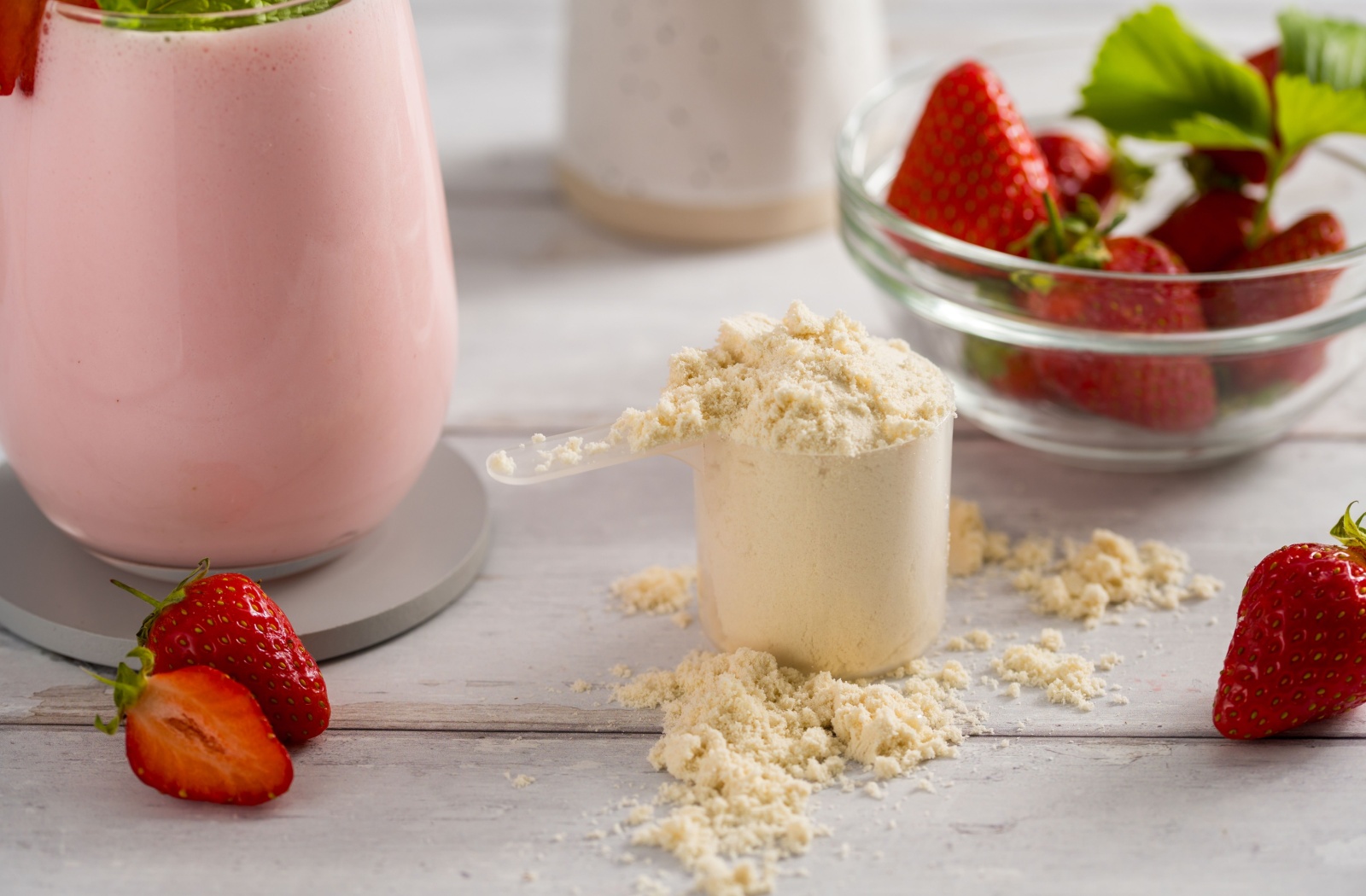 Close up of a scoop of protein powder next to a bowl of strawberries and a strawberry smoothie.